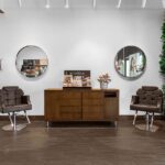 Modern salon interior with two brown chairs, a wooden cabinet displaying makeup products, two round mirrors, and a green vertical plant wall on the right. - Salon Dulay, FL