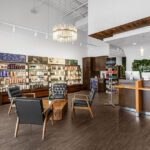 Modern salon reception area with wooden floors, product shelves, a chandelier, three chairs around a small table, and a front desk with two computers. - Salon Dulay, FL