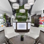 Modern salon reception area with white chairs, a small table, illuminated front desk, and shelves displaying hair and skincare products. Greenery and large hanging lights decorate the space. - Salon Dulay, FL