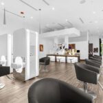 Modern hair salon with white partitioned styling stations, black salon chairs, wood flooring, and bright overhead lighting. The space appears clean and spacious with large windows at the back. - Salon Dulay, FL