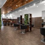 Modern hair salon interior with black styling chairs, large mirrors, wood flooring, green wall plants, and bright ceiling lights. - Salon Dulay, FL