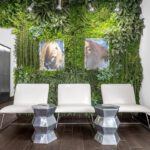 Three white chairs and two metallic tables are arranged in front of a green plant wall with two framed photos in a modern waiting area. - Salon Dulay, FL