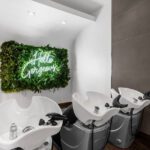 Three white salon shampoo stations are positioned beside a green wall with a neon sign that reads "Hello Gorgeous. - Salon Dulay, FL