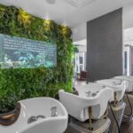 Row of white salon washbasins facing a lush green plant wall with a mission statement plaque; modern, well-lit interior. - Salon Dulay, FL