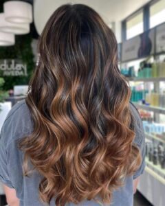 A person with long wavy hair featuring dark roots and caramel brown balayage highlights stands in a salon, facing away from the camera. - Salon Dulay, FL