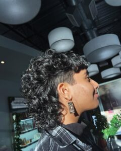 Person with a curly mullet hairstyle and shaved sides, wearing patterned earrings and a plaid shirt, standing indoors under round ceiling lights. - Salon Dulay, FL