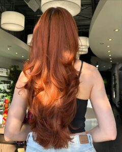 A person with long, wavy, copper-brown hair stands indoors, facing away from the camera, wearing a black top and light blue jeans. - Salon Dulay, FL