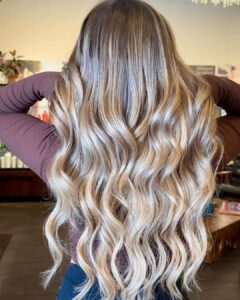 Person with long, wavy, blonde balayage hair seen from behind, wearing a brown long-sleeve shirt inside a salon. - Salon Dulay, FL