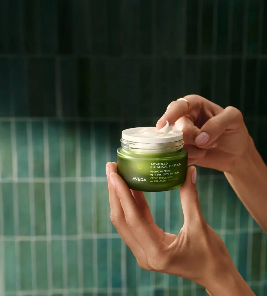 A person holds and opens a green jar of Aveda face cream against a tiled green background—perfect for spring prep to refresh your look. - Salon Dulay, FL