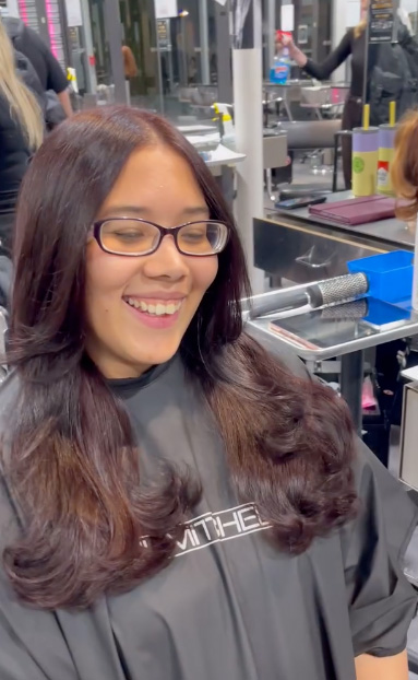 A woman with long, wavy brown hair and glasses smiles while sitting in a Salon Dulay chair in Orlando, wearing a black salon cape, ready to enjoy natural hair services. - Salon Dulay, FL
