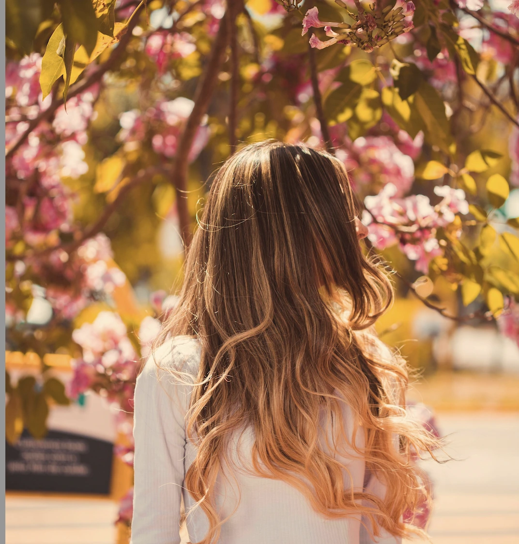 A person with long, wavy, blonde hair stands outdoors facing away, surrounded by blooming pink flowers and green leaves in sunlight. - Salon Dulay, FL