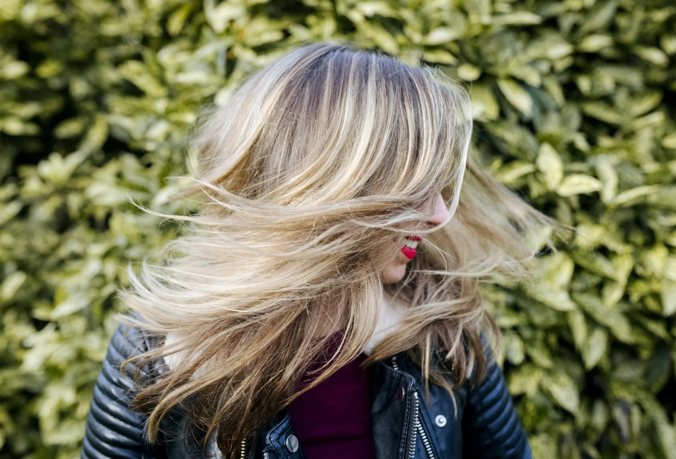 A person with long, blonde hair wearing a black leather jacket turns their head, causing their hair to fan out, with green foliage in the background. - Salon Dulay, FL